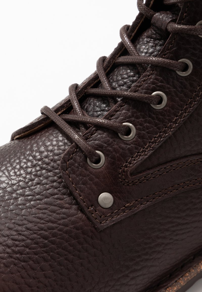 Braune Lederstiefel mit strukturiertem Obermaterial, Metallösen und kontrastierenden Nähten. Die Schnürsenkel sind durch die Ösen geflochten und sorgen für einen sicheren Sitz.