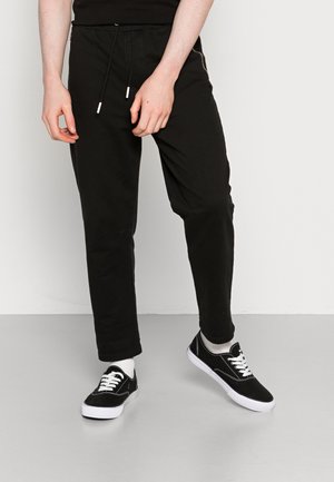 Person, die schwarze Kordelzughosen mit weißen Socken und schwarzen Turnschuhen trägt, steht vor einem schlichten weißen Hintergrund.