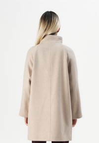 Femme aux longs cheveux blonds portant un manteau en laine beige avec un col montant, vue de dos sur un fond blanc uni.