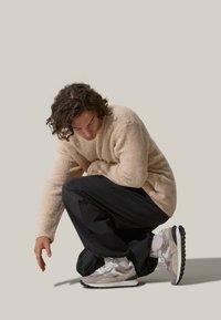 Maglione beige peloso, pantaloni neri a fit morbido e sneakers atletiche grigie con materiali misti e accenti texturizzati. Posizione inginocchiata su uno sfondo chiaro.