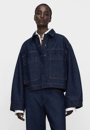 Dunkelhäutige Frau trägt übergroße dunkelblaue Jeansjacke und passende Hose, weiße Hemdärmel sind an den Ärmeln sichtbar.