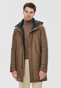 Brązowa ocieplana kurtka z kapturem, szary podkolanówka, zamek błyskawiczny z przodu oraz napy. Połączona z beżowym golfem i beżowymi spodniami.