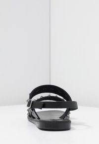 Sandalia de cuero negra con suela gruesa, correa ajustable para el tobillo y detalles de tachas en la parte trasera. Diseño minimalista con estructura abierta.