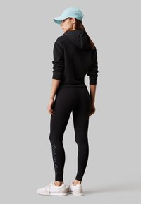 Vrouw draagt een zwarte hoodie, zwarte Calvin Klein leggings, witte sneakers en een lichtblauwe pet, staand tegen een grijze achtergrond.