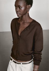 Cardigan marrone lavorato a maglia con collo rotondo, chiusura frontale con bottoni e orlo a coste. Abbinato a pantaloni bianchi e una cintura con borchie. Accessori semplici in argento.