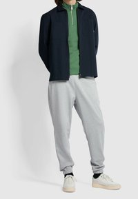 Marineblaue Jacke mit Reißverschluss über einem grünen hochgeschlossenen Pullover, kombiniert mit hellgrauen Jogginghosen und beigen Sneakers. Der Stoff wirkt weich und bequem.