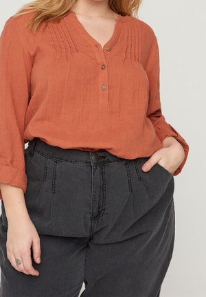 Frau trägt rostorangefarbene Bluse mit Knöpfen und Falten sowie hoch taillierte schwarze Hose, Hand in der Tasche, heller Hintergrund.