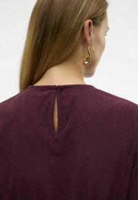 Burgundska bluzica iz teksturirane tkanine, z detajlom v obliki ključavnice na hrbtni strani, zaključena z okroglo ovratnikom. Zlati uhani v obliki spirale poudarjajo videz.