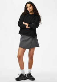 Femme aux longs cheveux bouclés, debout les bras croisés, portant un sweat à capuche noir, une jupe grise, des chaussettes blanches et des baskets noires sur un fond blanc.