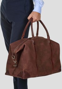 Mann i marineblå bukser og blå skjorte som holder en stor brun lær duffelbag i håndtakene mot en grå bakgrunn.