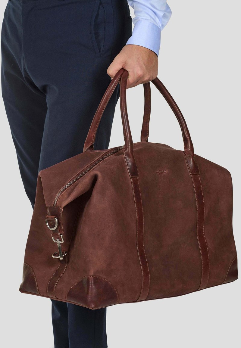 Mann i marineblå bukser og blå skjorte som holder en stor brun lær duffelbag i håndtakene mot en grå bakgrunn.