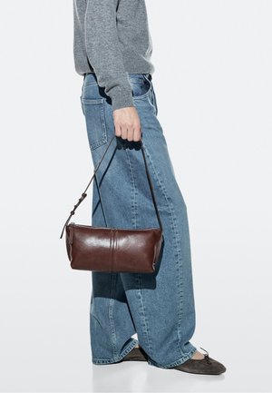 Persoon die een grijze trui en wijde blauwe spijkerbroek draagt, houdt een donkerbruine leren handtas bij het hengsel aan de zijkant vast.