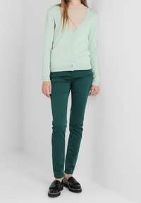 Cardigan de manga larga verde claro con escote en V y cierre de botones, combinado con pantalones ajustados verde oscuro y mocasines negros brillantes.