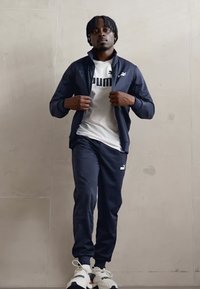 Jūras spēku treniņu tērps ar rāvējslēdzēju uz augšdaļas un pieguļošām biksēm, uz kreisā krūts un biksēm atrodas balts Puma logotips, apvienots ar baltām sporta apaviem.