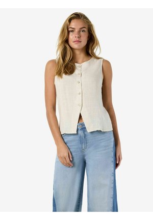 Femeie purtând un top bej fără mâneci, cu nasturi, asortat cu blugi albastru deschis cu picioare largi, stând în fața unui fundal alb simplu.