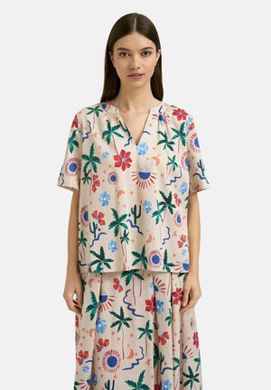 Frau trägt beige Kurzarmtop und Rock mit buntem Blumen-, Palmen-, Sonne- und Mondmuster, steht vor einfarbigem Hintergrund.