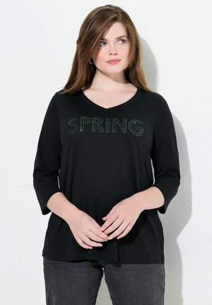 Zwarte langemouwen V-hals top met "SPRING" in groene rhinestones. Zachte stof met een relaxte pasvorm, gecombineerd met donkergrijze broek.