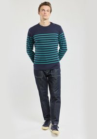 Pull rayé bleu marine avec des rayures turquoise, col rond, détail de bouton à l'épaule, associé à un jean en denim foncé et des baskets.