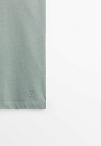 Partie inférieure d’un vêtement tricoté vert clair avec un petit logo circulaire brodé près de l’ourlet sur fond blanc.