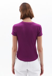 Kortærmet, tætsiddende lilla t-shirt i en blød, ribbet tekstur med rund halsudskæring og en buet kant, båret med hvide bukser.