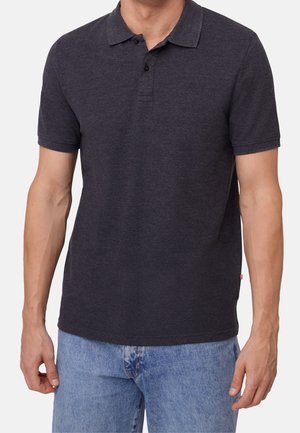 Mężczyzna ubrany w ciemnoszarą krótką koszulkę polo z dwoma guzikami i jasnoniebieskie dżinsy, stojący na jednolitym tle.