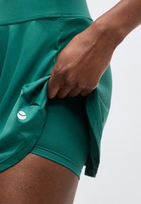 Ruka dvíha zelenú športovú sukňu, pod ktorou sú viditeľné priliehavé šortky so zodpovedajúcim dizajnom a malým bielym logom na sukni.