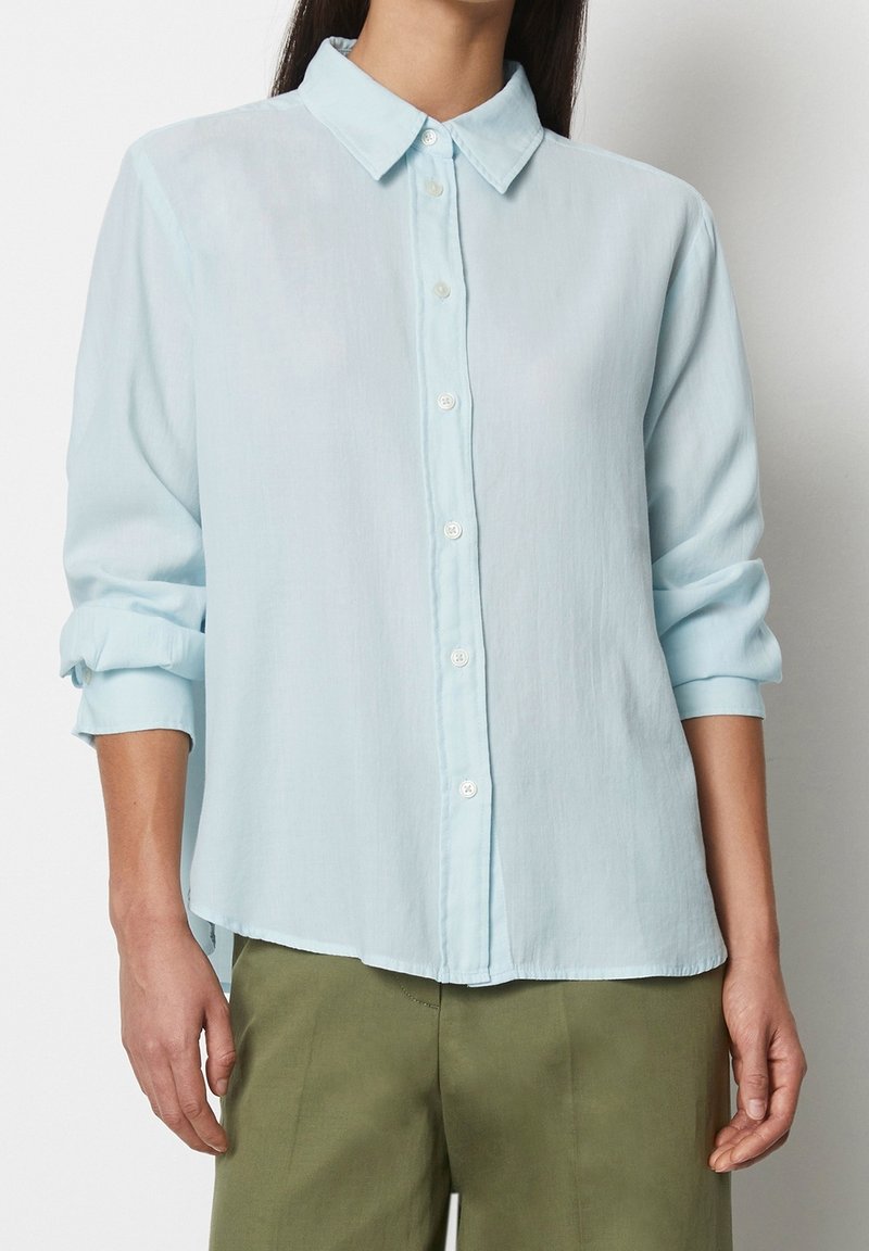 Personne portant une chemise boutonnée bleu clair à manches longues avec les manches retroussées et un pantalon vert olive, debout devant un fond uni.