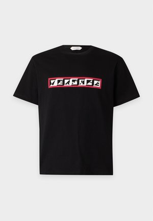 Homme portant un t-shirt noir Versace, pantalon ample gris à rayures verticales, chaussures noires et lunettes de soleil noires, debout avec une main dans la poche.