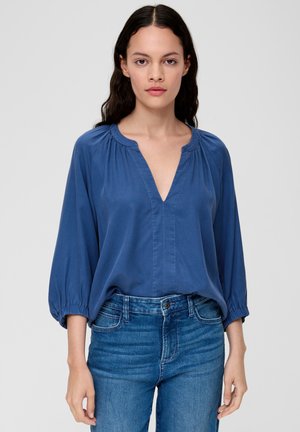 Blouse bleue, ample, avec un décolleté en V plongeant, des détails froncés au niveau du col et des manches élastiques. Associée à un jean en denim bleu.