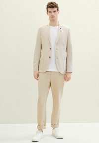 Blazer beige clair avec deux boutons et une poche à rabat, associé à un t-shirt blanc et un pantalon beige clair, complété par des baskets blanches.