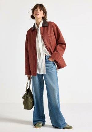 Donna che indossa una giacca rossa, una camicia bianca a righe, jeans larghi blu, scarpe verdi e tiene una borsa in camoscio verde.