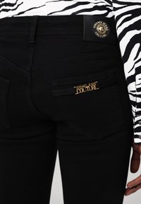 Jeans de mezclilla negros con un diseño ajustado, bolsillos traseros con herrajes en tono dorado "Versace Jeans Couture" y una etiqueta de piel en la cintura.