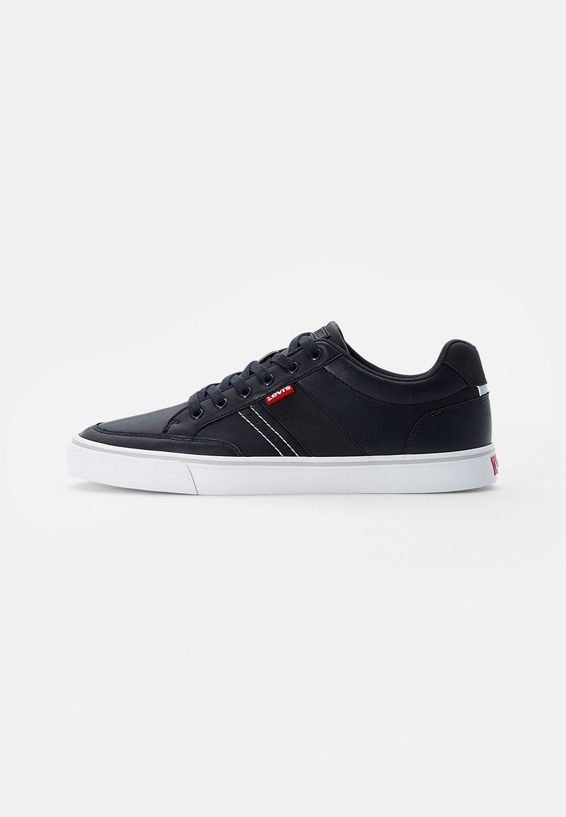 Tenis de couro preto com solas de borracha brancas, apresentando riscas brancas nas laterais e um pequeno logótipo vermelho na língua. Design minimalista.