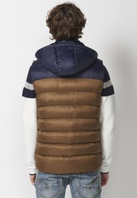 Padded jacket with a navy hood, brown body, and white striped sleeves. Features quilted texture and color block design.