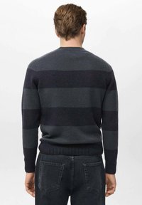 Uomo che indossa un maglione a righe blu scuro e grigio e jeans neri, in piedi di spalle contro uno sfondo chiaro e semplice.