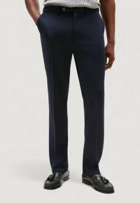 Pantaloni blu navy con gambe dritte, realizzati in tessuto liscio, dotati di passanti per cintura, bottone frontale e tasche laterali. Indossati con mocassini neri.