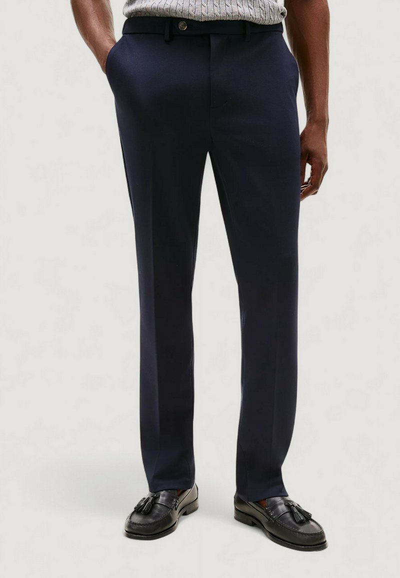 Pantaloni blu navy con gambe dritte, realizzati in tessuto liscio, dotati di passanti per cintura, bottone frontale e tasche laterali. Indossati con mocassini neri.