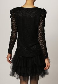Top de encaje negro con mangas largas y transparentes, y una falda de tul negro con volantes. La tela texturizada presenta un patrón floral.
