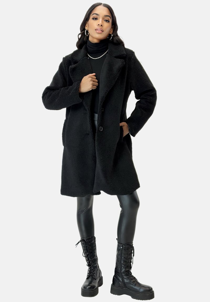 Black, fur-textured coat with a notched collar, single-button closure, large pockets, worn over fitted black top and leggings; paired with combat boots.