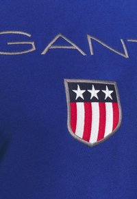 Camiseta de algodón azul con el logo de "GANT" bordado en plata y un parche con un escudo que presenta tres estrellas y franjas rojas y blancas en el pecho.