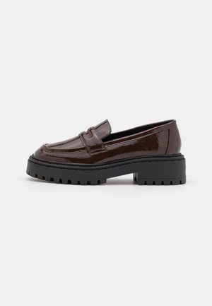 Mocassin marron foncé brillant avec semelle épaisse noire à crampons et petit talon carré, vue de côté sur fond blanc.