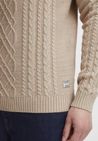 Beige strikkegenser med kabelmønster, ribbestrikket kant og mansjetter, samt en liten merkeetikett på siden. Strukturert, teksturert design.