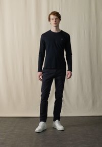 Sort langærmet skjorte parret med mørke bukser og hvide sneakers. Skjorten har et lille logo på brystet. Glat stof, tætsiddende design.