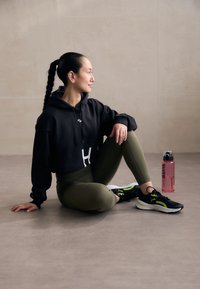 Felpa nera corta con scritte bianche, leggings verde oliva, scarpe da corsa nere con accenti neon e una bottiglia d'acqua rosa accanto.