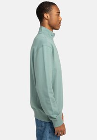 Green sweatshirt with a high collar, ribbed cuffs, and hem. Soft texture and loose fit, paired with light blue denim jeans.