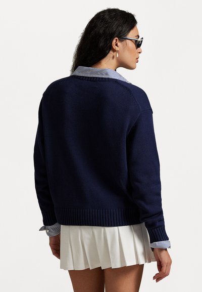 Navy blue knitted sweater with a ribbed hem and cuffs, layered over a white shirt with blue stripes, paired with a pleated white skirt.