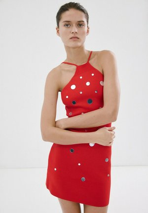 Joven mujer con un vestido sin mangas rojo adornado con círculos plateados, de pie con los brazos cruzados frente a un fondo blanco y liso.