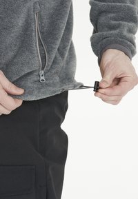 Pullover in pile grigio con tasca frontale con zip, dotato di un cordino all'orlo, abbinato a pantaloni neri, che cattura una mano mentre regola il cordino.
