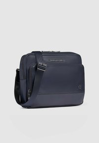 Borsa a tracolla in pelle blu navy con chiusura a zip, tracolla regolabile e dettaglio con logo. Texture liscia e forma rettangolare.