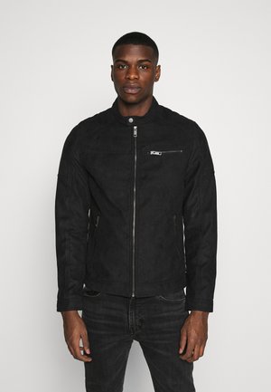 Chaqueta de ante negra con cremallera frontal, un bolsillo en el pecho con cremallera y cuello alto. Presenta detalles de costura y un diseño ajustado.